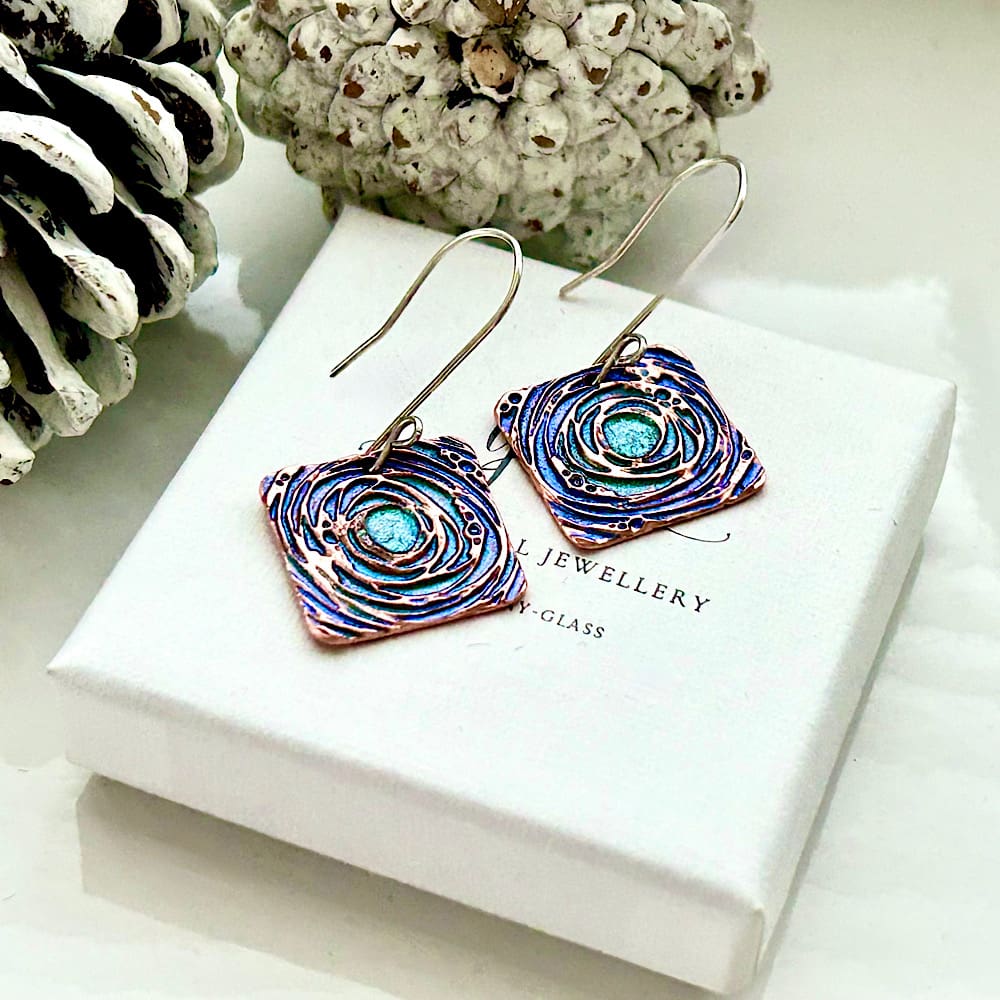 À pair of copper drop earrings in a square shape with a intucate pattern and hand painted in blue and Green metallic paints, they have sterling silver ear wires in a fish book shape. The earrings are displayed on a black velvet insert in a White gift box.