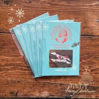 The picture shows a stack of five booklets titled 'A Wartime Christmas'. The booklet sits on a rich brown wooden table top which compliments the blue cover of the booklet. The cover shows the title text wrapped around a drawing of a laughing Santa, all in red. Underneath is a photo of the finished reusable fabric crackers, made to the pattern from 1940s wartime Britain.