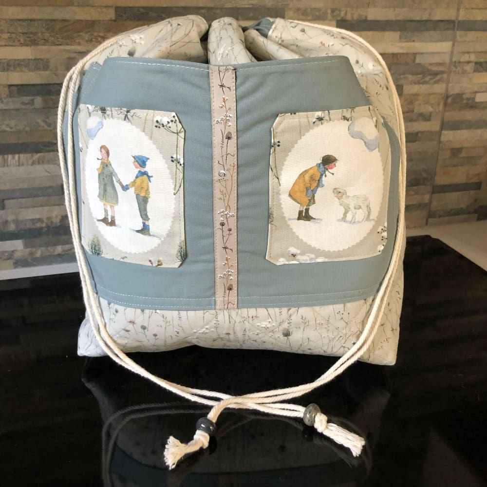 An extra large sized quilted drawstring bag in a grey blue fabric featuring winter grasses. Large front pocket features two children's shepherd figures. Closing using a natural cord secured by chunky blue grey ceramic beads.