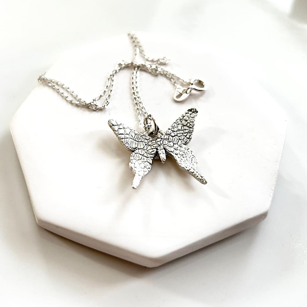 A fine silver pendant in the shape of a small butterfly, the pendant has a sterling silver chain. It is displayed on a black velvet pad inside a small white presentation box.