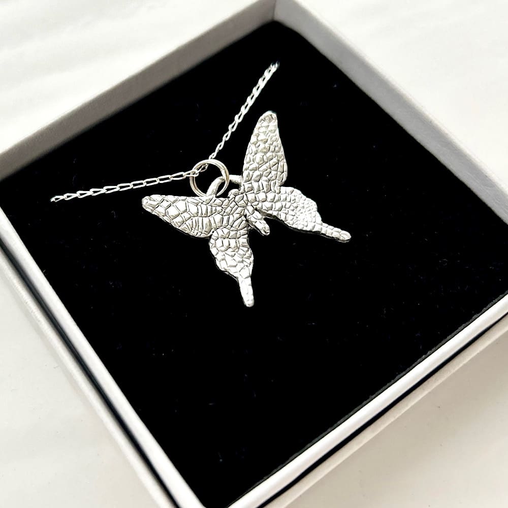 A fine silver pendant in the shape of a small butterfly, the pendant has a sterling silver chain. It is displayed on a black velvet pad inside a small white presentation box.