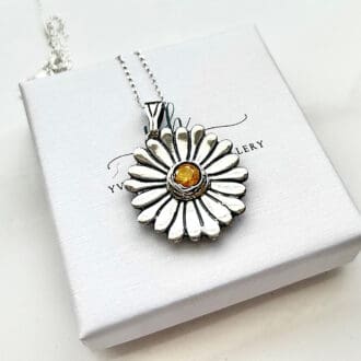 A fine silver pendant in the shape of a daisy flower with a yellow cubic zirconia in the centre. It has a sterling silver chain and is sat on a small white jewellery gift box.