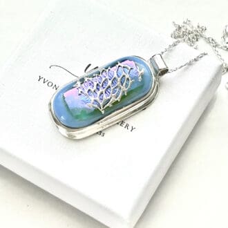 A blue fused glass oblong cabochon set in a fine silver frame with a fine silver lace pattern fused on top of the glass. The pendant has a sterling silver chain and is disolayed in a white presentation box with a black velvet base.