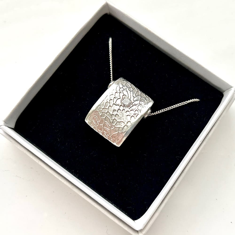 A fine silver double sided pendant with a small 4mm cubic zirconia stone set in the centre. It is patterened on both sides with plain silver edges and has a sterling silver chain, displayed in a white presentation box sat on black velvet.