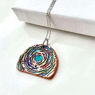 À half moon shaped copper pendant which has been hand-painted with metallic paints in a mix of colours. It has a rise gold plated fine chain and set on a white background with a white gift box in the picture.