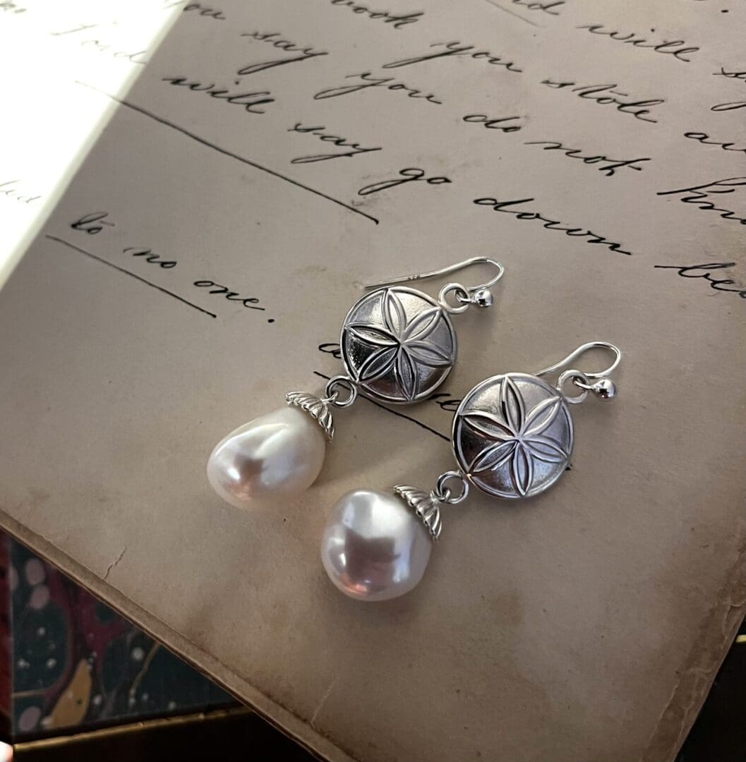 a pair of drop earrings in silver are laying on top of an open book. The earrings have a hook fitting, underneath which is a round hexafoil symbol and beneath there is an asymmetrical pearl in a shaped pearl cup