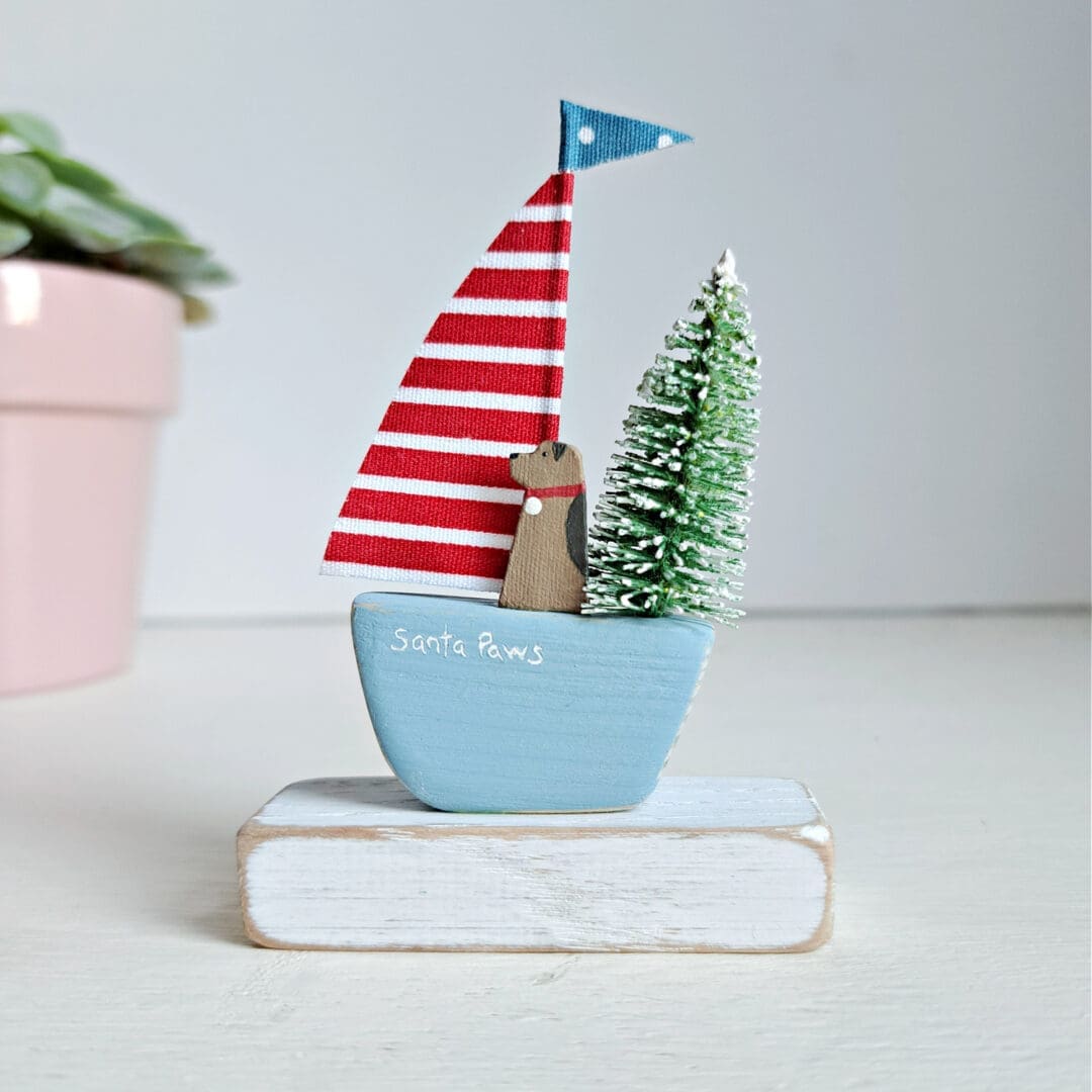 Sailing boat with a blue base and red and white stripe sail. A terrier dog is sitting in the boat with a christmas tree