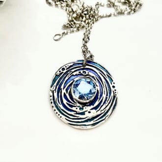 A handcrafted fine silver round shaped pendant that has a 8mm. blue cubic zirconia stone set in the centre. The pendant has been hand- painted in between the design pattern, in blue metallic points, the necklace has a sterling silver chain.