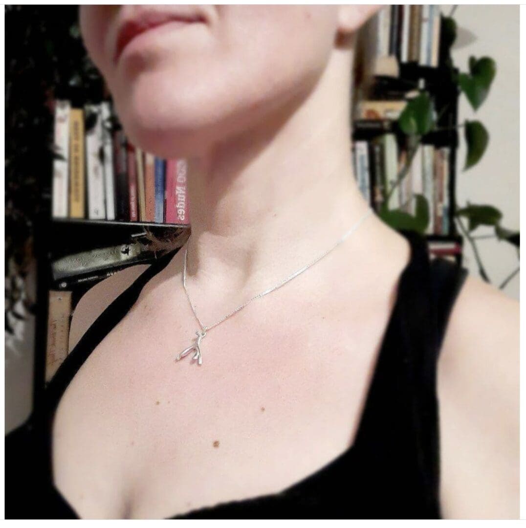 A person is wearing a small silver pendant in the shape of the internal clitoris around their neck. You can see a black sleeveless top and a bookshelf in the background