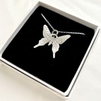 A fine silver pendant in the shape of a small butterfly, the pendant has a sterling silver chain. It is displayed on a black velvet pad inside a small white presentation box.