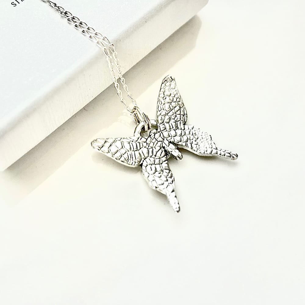 A fine silver pendant in the shape of a small butterfly, the pendant has a sterling silver chain. It is displayed on a black velvet pad inside a small white presentation box.