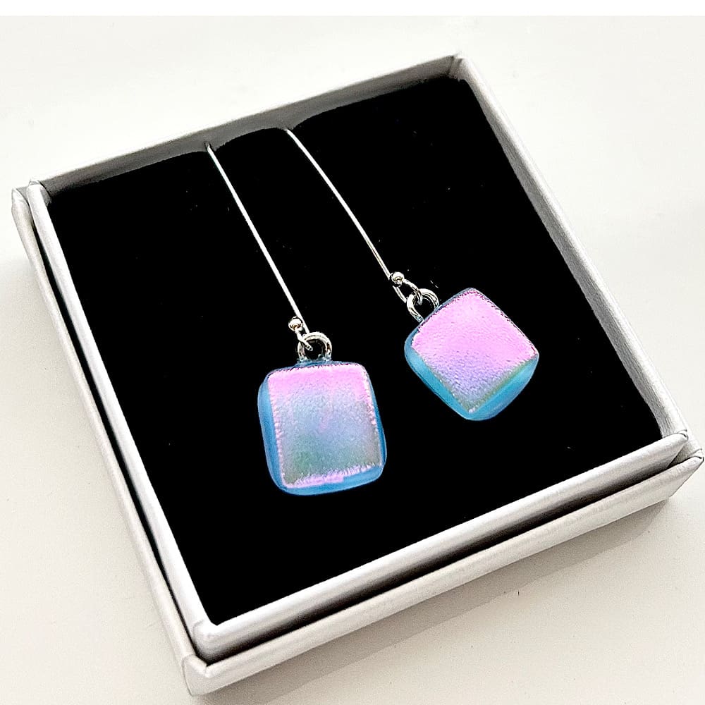 A pair of small fused glass long drop earrings with sterling silver ear wires. Dichroic glass in pale blue reflecting to pink with movement, a soft square shape, handcrafted