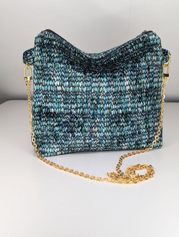 Knitted crossbody bag with a multicolored pattern in shades of blue, green, and black, featuring a gold chain strap on a white background