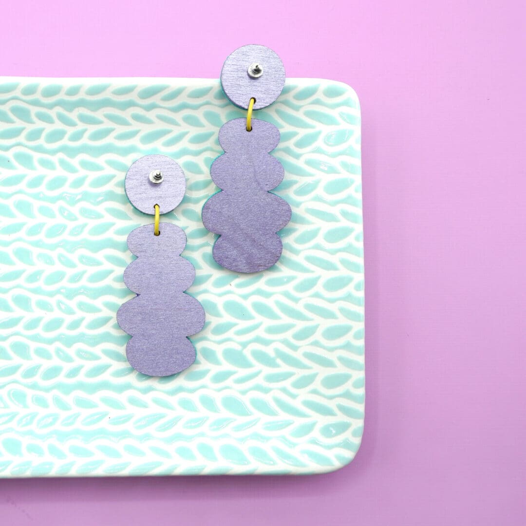 Colourful pastel geometric faux leather dangle earrings with abstract patterns and yellow connectors shown on a mint green dish against a pink backdrop.