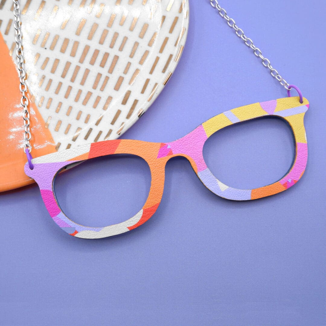 Colourful nerdy necklace shaped like spectacles shown against a denim blue coloured background