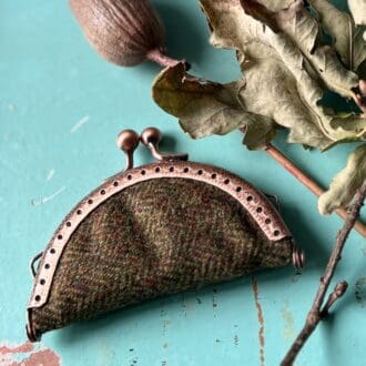 The picture shows a vintage style needle book laying on a blue painted wooden table, next to dried leaves and seed pods. The needle book is semi circular in shape and has been made using a metal purse clasp as its fastening.