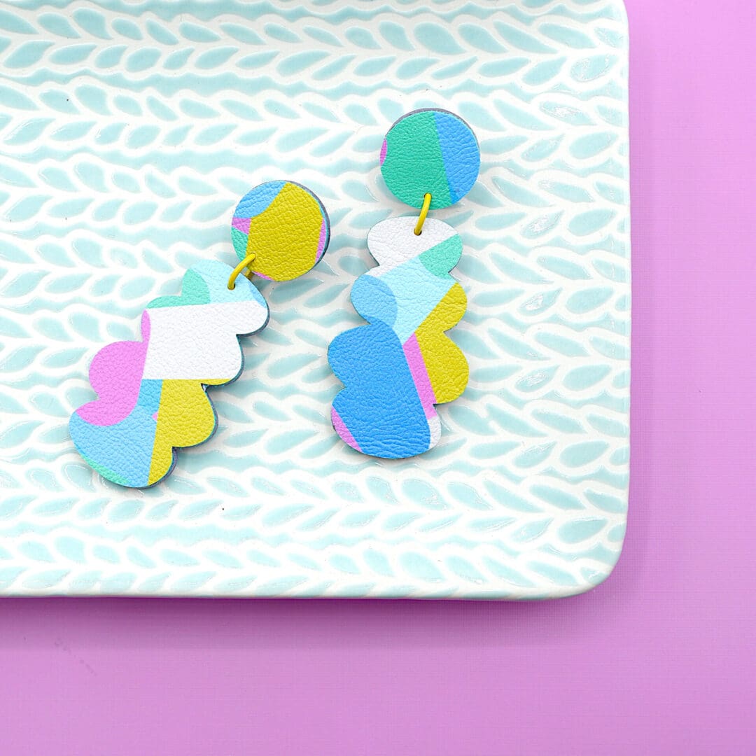 Colourful pastel geometric faux leather dangle earrings with abstract patterns and yellow connectors shown on a mint green dish against a pink backdrop.