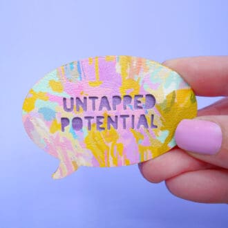 A quirky colourful speech bubble brooch is shown held in a white woman's hand. The badge is made from wood and faux leather printed with an original design. It's a textured abstract art pattern featuring golden yellows, dusky pink and mauve with touches or green and blue