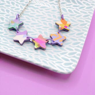 Colourful star necklace made from faux leather, featuring vibrant abstract patterns in pastel shades of purple, orange, yellow, and pink. The stars are linked together with purple jump rings and displayed on a textured pale blue dish, against a pink background.