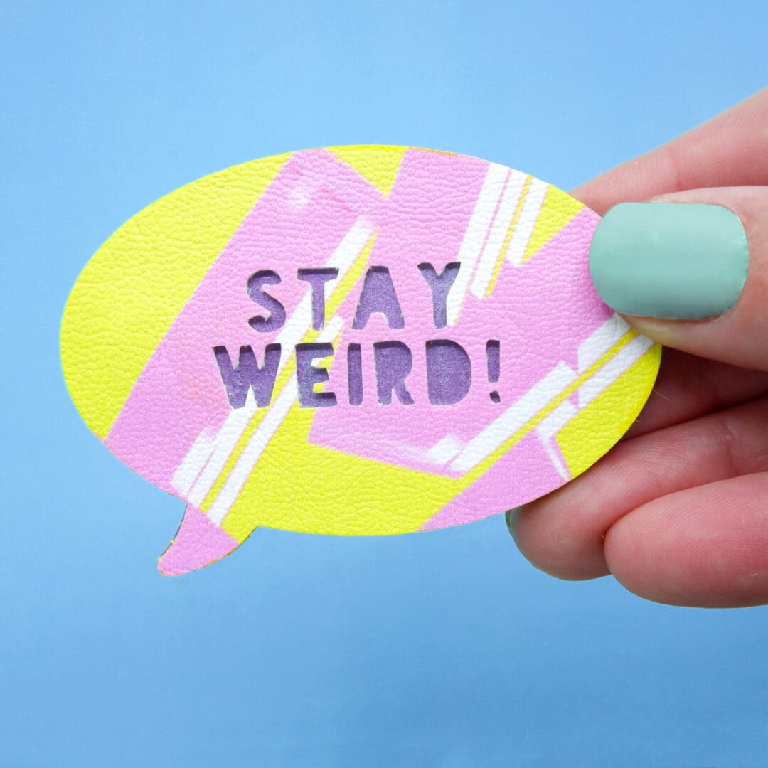 Close-up of a colourful faux leather brooch in the shape of a speech bubble with the words 'Stay Weird!' laser-cut into it, held against a bright mint green background.