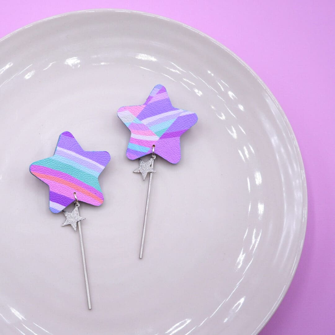 Pair of star-shaped earrings with pastel purple, pink, turquoise, and white stripes. Each earring features a glittery silver star charm and a long silver stick, set on a soft white plate with a pink background.