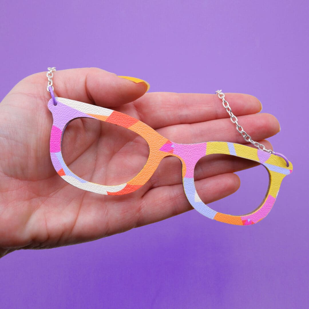 Bold bright coloured necklace shaped like a pair of glasses. Shown held in white woman's hand against purple background