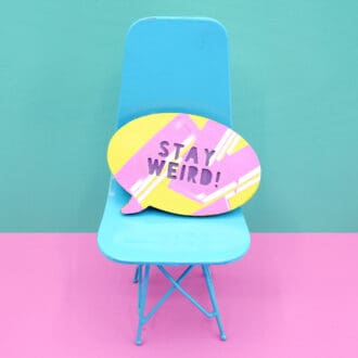 "A vibrant faux leather brooch with 'Stay Weird!' text sits on a miniature turquoise chair against a pink and mint green background, showcasing its playful and quirky design."