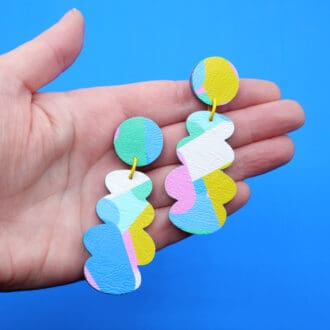 A pair of colourful drop stud earrings made from faux leather, featuring pastel abstract patterns in pink, blue, green, white, and yellow, held in a hand against a vibrant blue background.