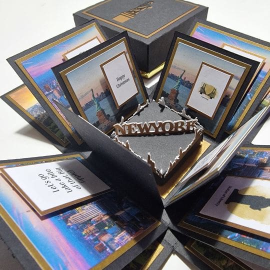 a stunning pop-up box card, elegantly themed around New York City. When opened, it reveals an array of beautifully decorated panels. The central feature is a bold 3D cutout of the word "NEW YORK." Surrounding this centerpiece are panels showcasing the iconic New York skyline and festive messages like "Happy Christmas" and "Let's go on a date to the top of the Empire State Building." It's an imaginative and eye-catching design, making it a perfect choice for a special celebration