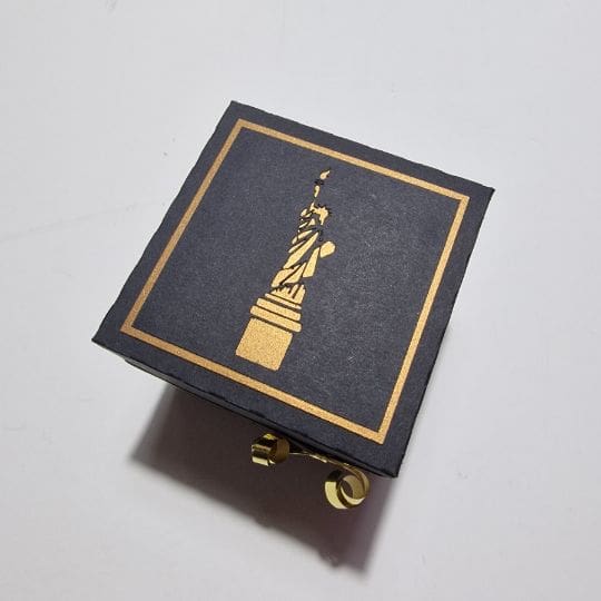 The image depicts a black box with a golden outline of the Statue of Liberty on its top surface. The box appears to be made of paper or cardboard and has a simple yet elegant design. The golden Statue of Liberty stands out against the black background, creating a minimalist and artistic presentation.