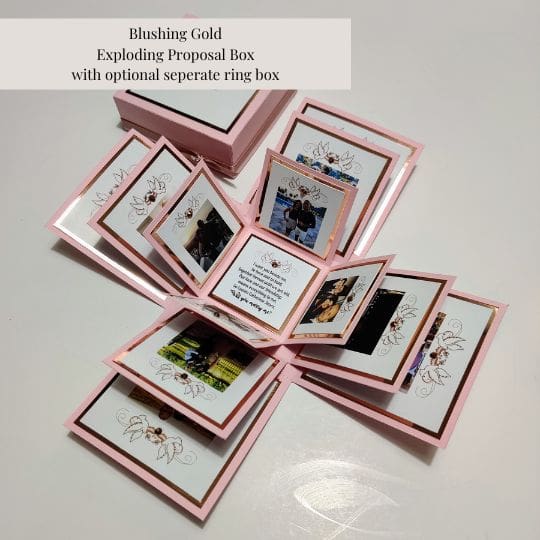 The image shows a 'Blushing Gold Exploding Proposal Box with optional separate ring box.' The box is pink with gold accents and opens up to reveal multiple layers of smaller cards or compartments. Each compartment contains photos and decorative elements, making it a visually appealing and creative way to present a proposal. The design is intricate and personalized, which makes it interesting and relevant for special occasions like proposals.