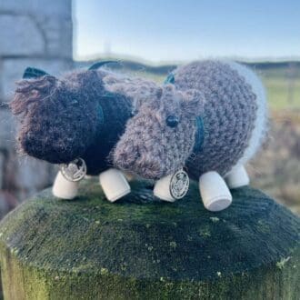 Two crocheted Bo' Beltie Galloway cows sit on top of a wooden post. Dark brown and light brown with a white stripe around their stomachs. Tartan ribbon around the necks with a silver Scottish thistle charm. Wooden legs. Design by Midge Porter Design. Design of a cow breed from Scotland.