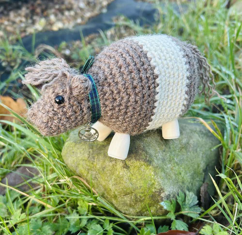 A crocheted Bo' Beltie Galloway cow sit on top of a grassy stone. Light brown with a white stripe around the stomach. Tartan ribbon around the necks with a silver Scottish thistle charm. Wooden legs. Design by Midge Porter. Design of a cow breed from Scotland.