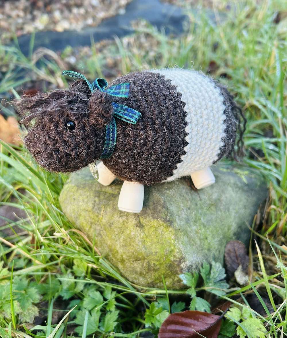 A crocheted Bo' Beltie Galloway cow sit on top of a grassy stone. Dark brown with a white stripe around the stomach. Tartan ribbon around the necks with a silver Scottish thistle charm. Wooden legs. Design by Midge Porter. Design of a cow breed from Scotland.