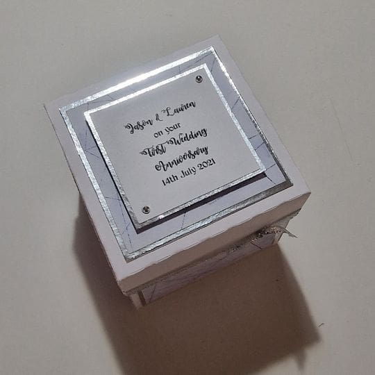 A small, square, white box with a decorative silver border. On the top of the box, there is a square card with a silver border and text that reads: "Jason & Laura on your 25th Wedding Anniversary 14th July 2021."