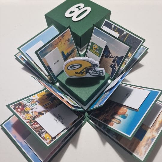 The image shows a pop-up card or box that has been opened to reveal multiple panels with images and text. The central feature is a 3D cutout of a football helmet with the Green Bay Packers logo. The box is green with the number '60' on the top, possibly indicating a 60th birthday or anniversary. Each panel contains various images, likely related to the Green Bay Packers, and some text.