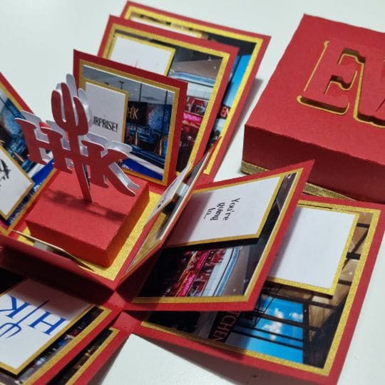 The image shows a red and gold pop-up card or box with multiple panels that open outward. The panels contain various images and text. In the center, there is a 3D pop-up element with the letters 'HK' and a trident-like symbol. The box lid, placed to the side, has the letters 'E' and 'K' cut out on it. The text on one of the panels reads, 'You're going to H.K