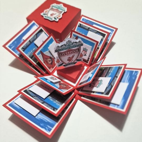 The image shows an intricately designed pop-up card or box featuring the Liverpool Football Club logo. The box is red with the Liverpool FC crest on the lid, and when opened, it reveals multiple layers of folded paper that expand outward. Each layer contains various images and text related to Liverpool FC, including the club's crest prominently displayed in the center. This design showcases a creative and detailed way to celebrate or present memorabilia for fans of Liverpool FC