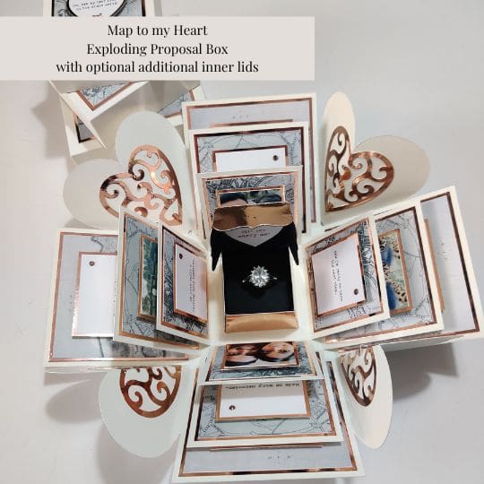 The image shows an 'Exploding Proposal Box' titled 'Map to my Heart' with optional additional inner lids. The box is open, revealing multiple layers of decorative panels and compartments. At the center of the box is a ring, suggesting that this is a proposal box. The panels are adorned with intricate designs, including heart shapes and framed sections that appear to hold messages or photos. This type of box is interesting and relevant as it provides a creative and memorable way to propose, combining elements of surprise and personalization