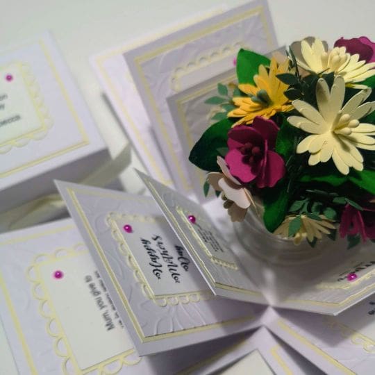 A pop-up card featuring a bouquet of paper flowers in the center. The card is open and reveals multiple panels with decorative elements and text. The flowers are detailed and colorful, with white, yellow, and pink petals, and green leaves. The card is handcrafted and designed for a special occasion like a birthday, anniversary, or celebration