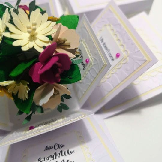 A beautifully crafted pop-up card displaying a bouquet of colorful paper flowers at the center. The card is open, revealing multiple intricately designed panels with various decorative elements and heartfelt text. The flowers, with their white, yellow, and pink petals, are complemented by green leaves, all carefully detailed to celebrate a special occasion