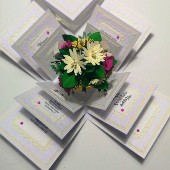 A beautifully crafted pop-up card displaying a bouquet of colorful paper flowers at the center. The card is open, revealing multiple intricately designed panels with various decorative elements and heartfelt text. The flowers, with their white, yellow, and pink petals, are complemented by green leaves, all carefully detailed to celebrate a special occasion