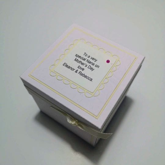 A small, light-colored gift box with a decorative label on top. The label has a scalloped border and a heartfelt message: 'To a very special Nana on Mother's Day, love, Eleanor & Rebecca.' The box is simple yet elegant, perfect for a cherished gift.