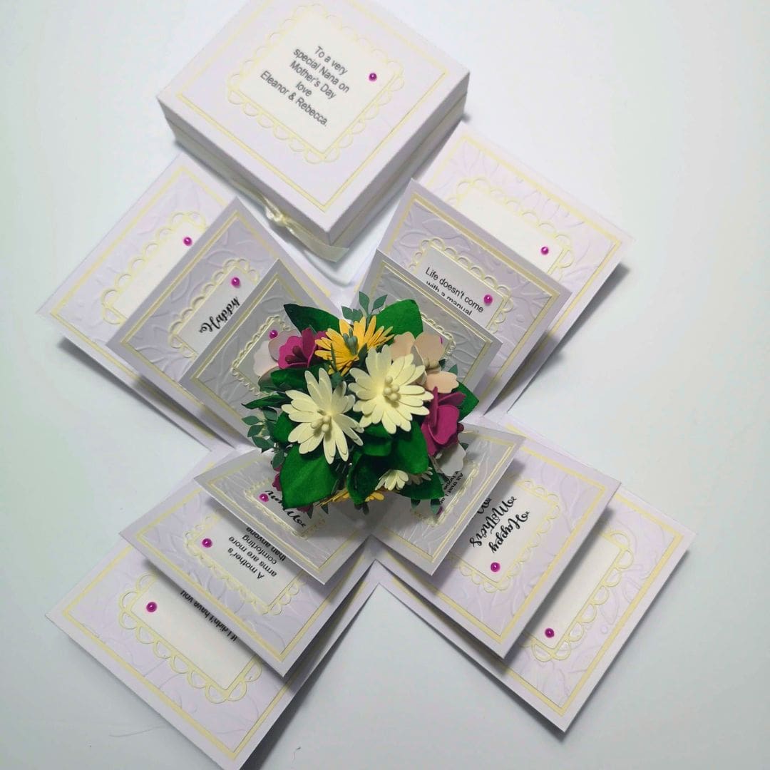 A pop-up card celebrating Mother's Day, featuring a bouquet of paper flowers at the center. The card is intricately designed with multiple layers of folded paper, decorative elements, and heartfelt messages. The outermost layer includes pink embellishments and panels with text, such as 'To a very special Mother’s Day on 14th May for Eleazar & Rebeccah,' 'Life doesn’t come with a manual, it comes with a mother,' 'Happy,' 'Beautiful Mother,' and 'You mean the world to me and I love you so much.