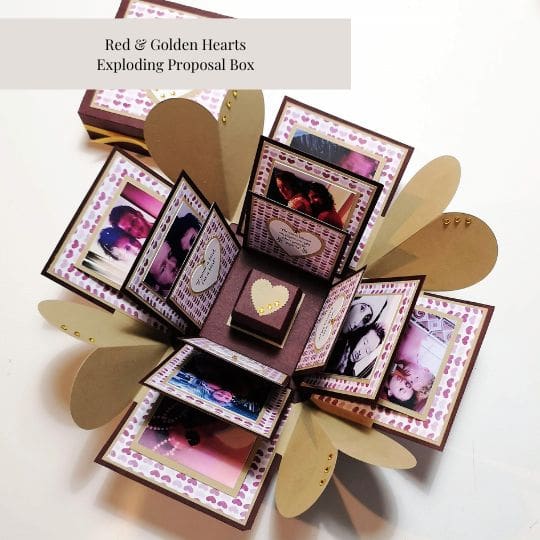 The image shows an 'Exploding Proposal Box' titled 'Red & Golden Hearts Exploding Proposal Box.' The box is open, revealing multiple layers of flaps decorated with photos and heart-shaped designs. The central part of the box contains a small box with a heart on top. The outer flaps are adorned with golden heart shapes, and the inner flaps display various photos and messages. This type of box is often used for special occasions like proposals, as it provides a creative and personalized way to present a surprise