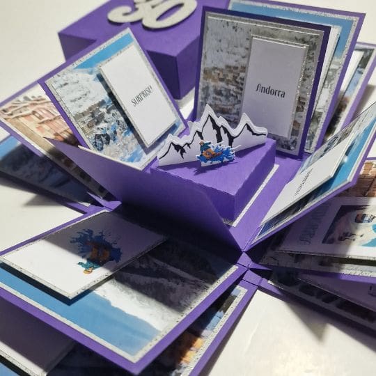 The image shows a purple pop-up card or box that has been opened to reveal multiple layers and sections. Each section contains various images and text related to snowy landscapes and locations. The central part of the card features a pop-up of mountains with a small figure skiing or snowboarding. The text visible in the image includes 'Andorra' and 'Switzerland.' The card appears to be a creative and intricate design, possibly for a special occasion or as a travel-themed gift