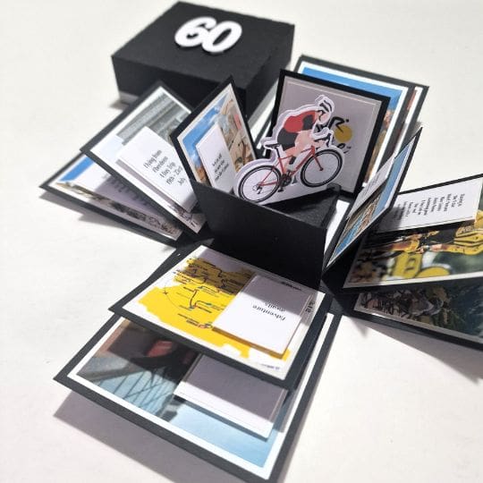 The image shows an open black box with the number '60' on the lid. The box is designed as an explosion box, which opens up to reveal multiple layers of folded paper and images. At the center of the box, there is a cutout of a cyclist on a bicycle. The surrounding layers contain various images and text, including a map and other pictures, possibly related to cycling or a travel theme. This type of box is often used for personalized gifts or special occasions, making it interesting and relevant for celebrations or commemorations