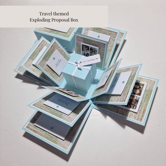 The image shows a 'Travel themed Exploding Proposal Box.' The box is light blue and opens up to reveal multiple layers of cards and photos arranged in a star-like pattern. Each layer contains various travel-themed decorations, messages, and photos, with a small gift box in the center. This type of box is interesting and relevant as it is a creative and personalized way to propose, incorporating travel memories and significant moments shared by the couple