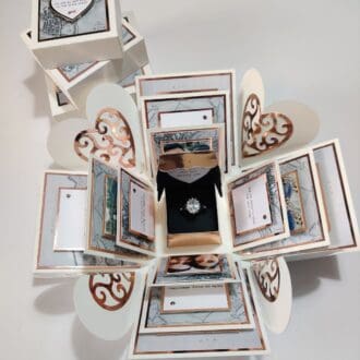 The image shows an intricately designed pop-up box that opens to reveal a ring in the center. The box has multiple layers with various decorative elements, including heart shapes and framed sections with text. The central compartment contains a ring, suggesting that this is a proposal box. The text in the center reads, 'Will you marry me?' This image depicts a creative and personalized way to propose marriage