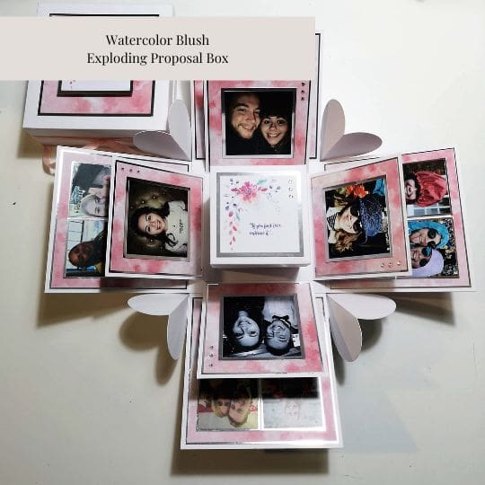 The image shows a 'Watercolor Blush Exploding Proposal Box.' The box is open, revealing multiple layers of photographs and decorative elements arranged in a way that they 'explode' outward when the box is opened. The central part of the box contains a smaller box with a floral design and text. The surrounding panels feature various photographs of people, arranged in a visually appealing manner with a watercolor blush theme. This type of box is often used for special occasions such as proposals, anniversaries, or other significant events, making it a unique and personalized gift